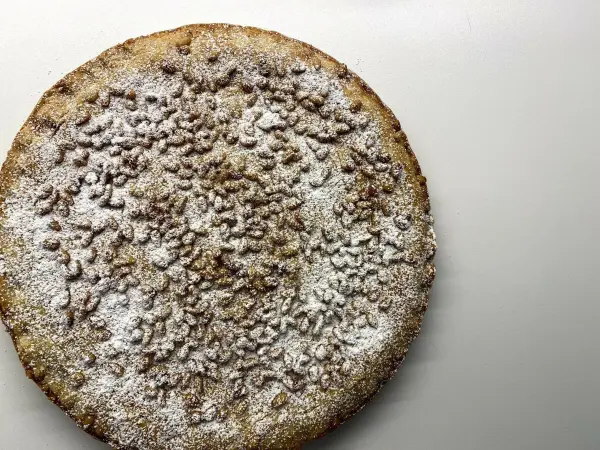 Ein runder Torta della Nonna liegt auf einer weißen Fläche, mit Puderzucker und Pinienkernen dekoriert.