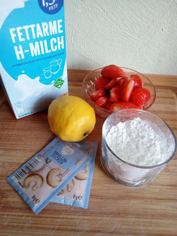 Ein Karton fettarme H-Milch und Zutaten wie Zitrone, Erdbeeren, Vanillinzucker sowie Puddingpulver auf einem Holzbrett.