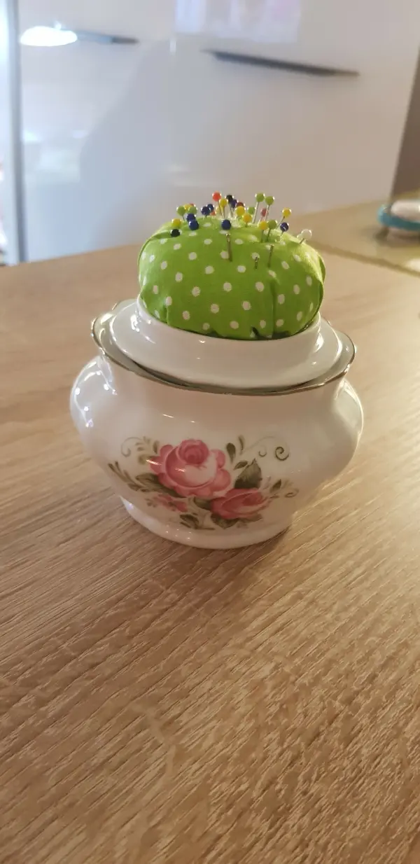Ein Keramikgefäß mit Rosenmuster hält ein grünes Nadelkissen, das mit bunten Stecknadeln dekoriert ist.