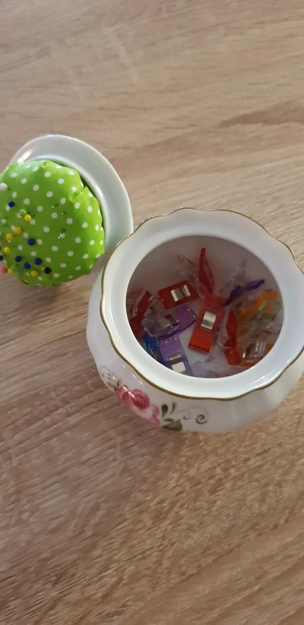 Ein Keramikgefäß mit einem bunten Nadelkissen auf dem Deckel enthält bunte Stoffclips und liegt auf einem Holzuntergrund.