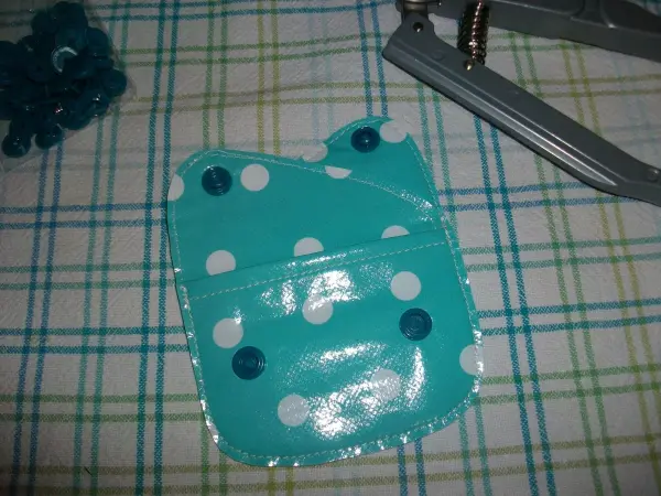 Eine farbenfrohe, gepunktete Geldtasche liegt auf einem karierten Tisch, während Druckknöpfe und ein Werkzeug daneben bereitliegen.