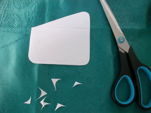 Eine Papierform für eine Geldtasche liegt auf einem grünen Tisch, während daneben Schere und ausgeschnittene Stoffreste liegen.
