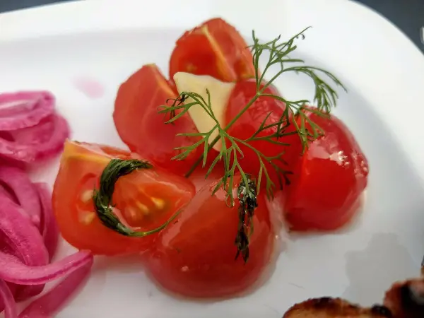 Eingelegte Tomaten liegen glänzend auf einem Teller, garniert mit frischem Dill, daneben pinke Zwiebeln.