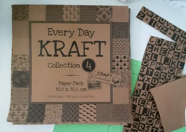 Ein Bastelpapierpaket mit verschiedenen Designs liegt auf einem Tisch, umgeben von Buchstabenschnipseln zur Kartengestaltung.