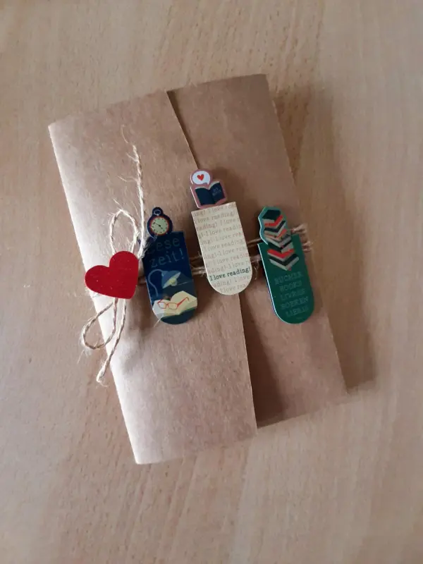 Eine dekorative Karte aus braunem Papier, liebevoll verziert mit Lesezeichen und einem roten Herz, liegt auf einem Tisch. Transkription: "Diese Zeit! I love reading! Bücher leben."