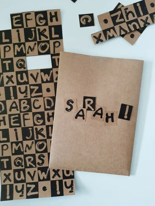 Eine handgemachte Glückwunschkarte mit dem Namen "SARAH!" steht auf einem Tisch, umgeben von Buchstabenschnipseln.