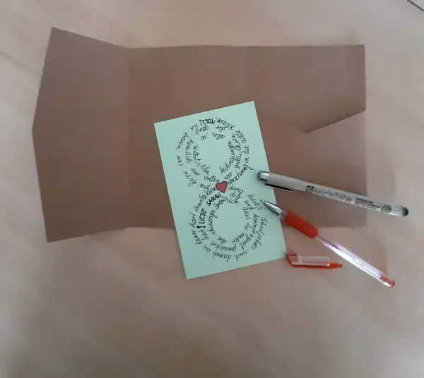 Eine grüne Glückwunschkarte zur Versetzung liegt auf dem Tisch, umgeben von Stiften und einem braunen Umschlag.