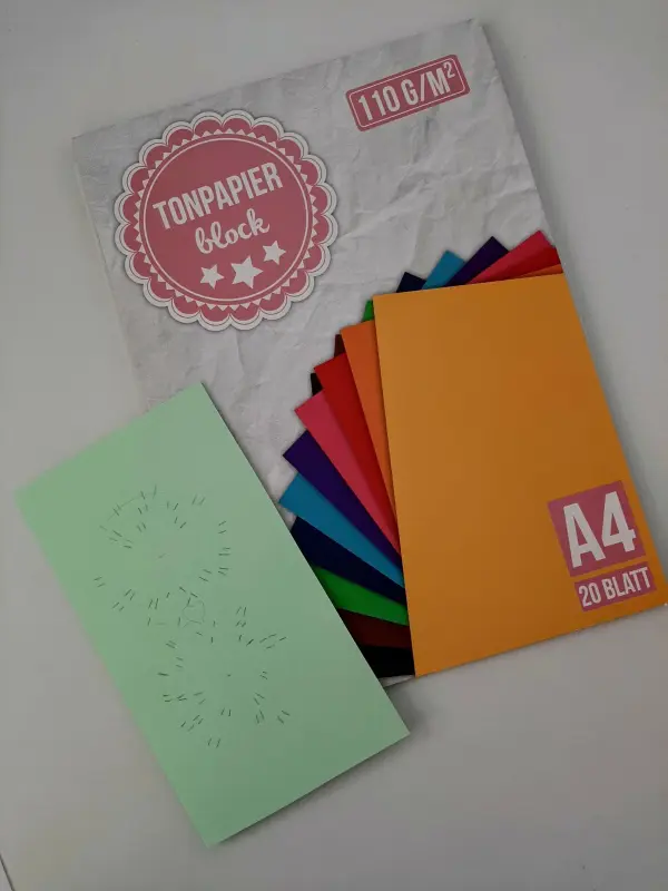Ein grünes Blatt mit einer dekorativen Stanzung liegt neben einem Block farbiger Tonpapiere, bereit für eine Glückwunschkarte zur Versetzung.