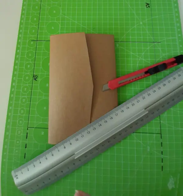 Ein braunes Kuvert liegt auf einem grünen Schneideunterlage, während ein Cutter und ein Lineal zur Vorbereitung für eine Glückwunschkarte verwendet werden.