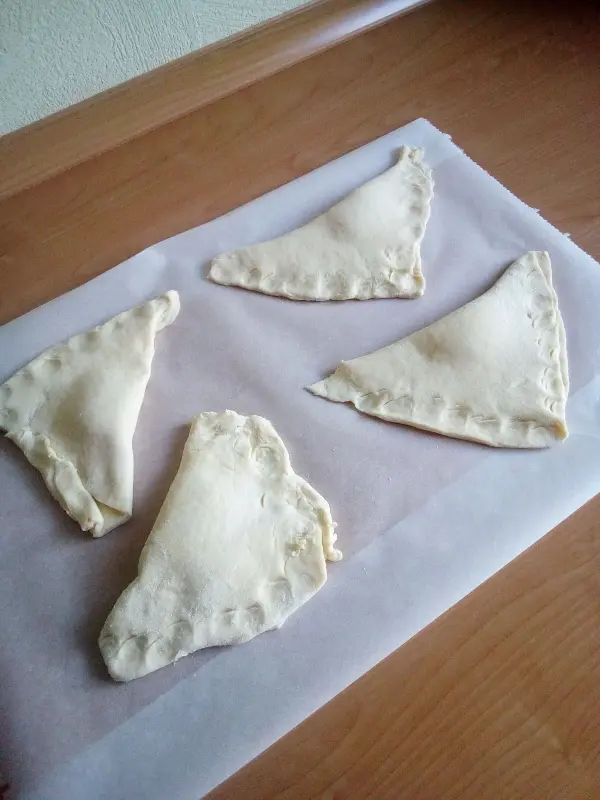 Viereckige Quark-Kissen aus Teig liegen auf Backpapier auf einer Holzoberfläche, bereit zum Backen oder Füllen.