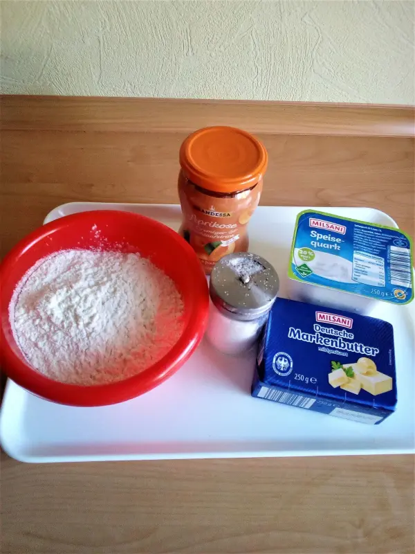 In einer roten Schüssel liegt Mehl, daneben Quark, Butter, Zucker und Aprikosenmarmelade auf einem weißen Tablett.