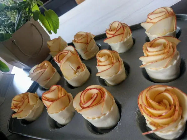 Zarte Apfelrosen aus dünn geschnittenem Apfel, in einem Muffinblech platziert, vor einem neutralen Hintergrund.