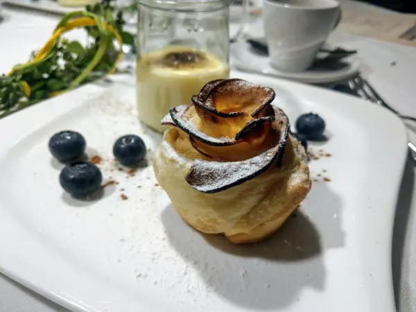Ein zartes Dessert in Form einer Apfelrose aus Blätterteig liegt auf dem Teller, umgeben von Heidelbeeren und Sahne.