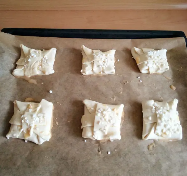 Sechs süße Blätterteig-Quark-Teilchen liegen auf einem Backblech, bereit zum Backen in einer Küche.