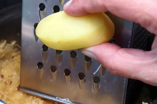 Eine Hand reibt eine Kartoffel auf einer Reibe über einer Schüssel mit Sauerkraut, um das Gericht schmackhafter zu machen.