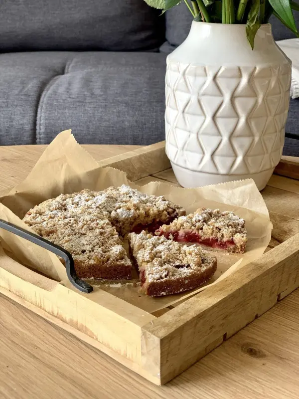 Ein frischer Streuselkuchen mit Obst liegt auf einem Holztablett, während eine Pflanze in einem dekorativen Topf daneben steht.