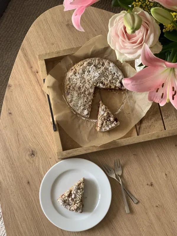 Ein goldbraun gebackener Streuselkuchen liegt auf einem Tablett, umgeben von Blumen; ein Stück wurde bereits entnommen.