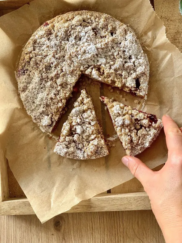Ein Streuselkuchen liegt auf Backpapier in einer Holzschale, während eine Hand ein Stück davon herausnimmt.