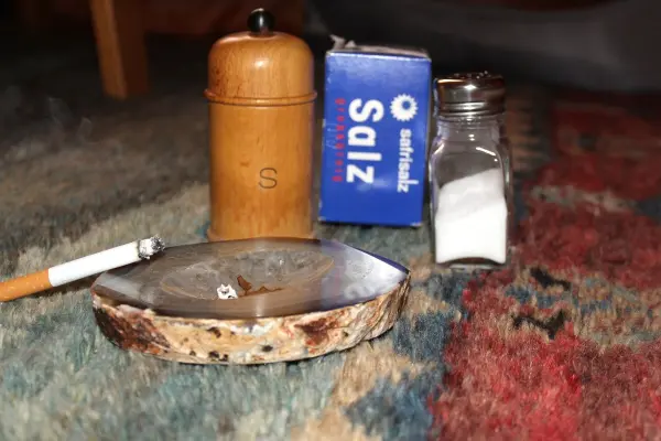Aschenbecher mit Zigarette und verbranntem Salz befindet sich auf einem bunten Teppich; daneben stehen Salzstreuer und -packung.