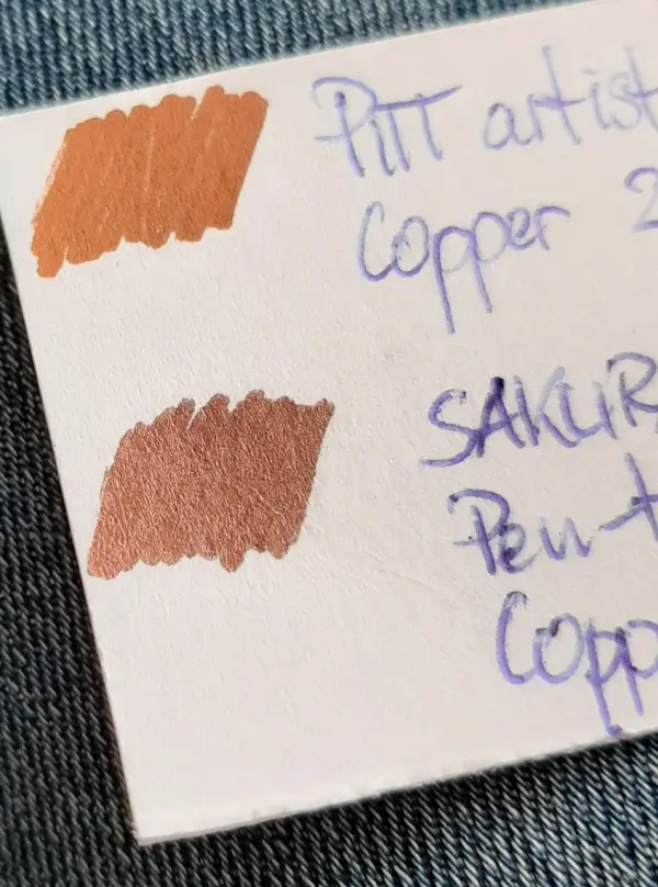 Beschrieben wird eine Farbpalette mit zwei gekritzelten Farbmustern in Kupfernuancen, auf weißem Papier vor blauer Jeans.