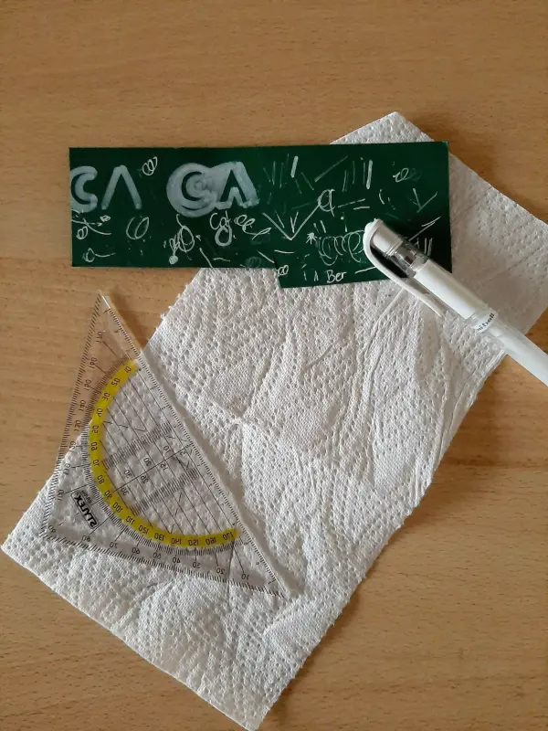 Ein gefalteter Tannenbaum liegt auf einem Tisch, umgeben von einem Geodreieck, einem Stift und Papiertüchern, die bei der Erstellung helfen.