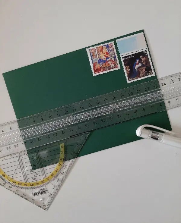 Ein gefalteter Tannenbaum aus grünem Papier liegt auf einem Tisch, umgeben von Lineal, Geodreieck und Stiften für die Gestaltung.