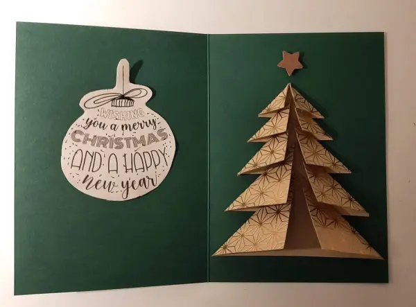 Eine gefaltete Weihnachtskarte zeigt einen goldfarbenen Tannenbaum, der festlich auf grünem Hintergrund steht. Oben hängt eine Weihnachtskugel mit dem Text: "Wishing you a merry Christmas and a happy new year".