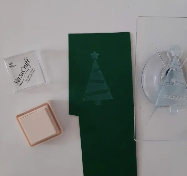Ein gefalteter Tannenbaum ist auf grünem Papier abgedrückt, umgeben von Stempelkissen und Stempelwerkzeug für kreative Weihnachtskarten.
