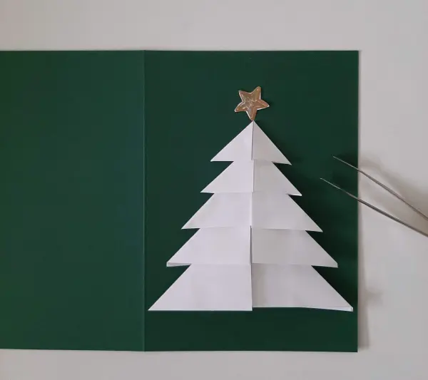 Ein gefalteter Tannenbaum aus weißem Papier steht auf einem grünen Hintergrund, verziert mit einem goldenen Stern oben.