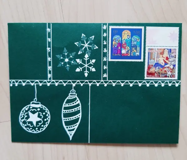 Eine grüne Weihnachtskarte zeigt einen gefalteten Tannenbaum und dekorative Elemente, mit Briefmarken und Schneeflocken.
