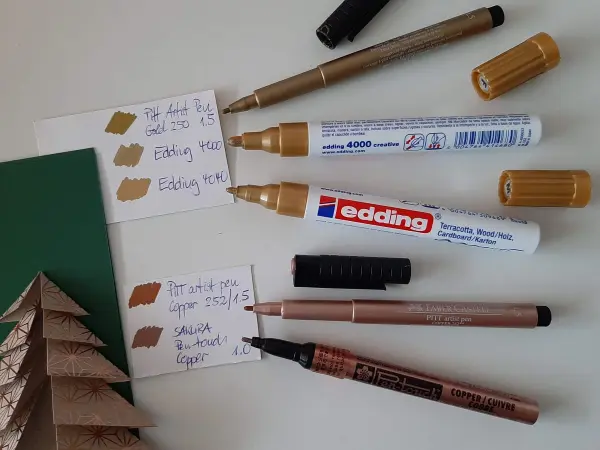 Ein gefalteter Tannenbaum liegt neben verschiedenen Markern und Farbproben auf einem Tisch, um Weihnachtskarten zu gestalten.