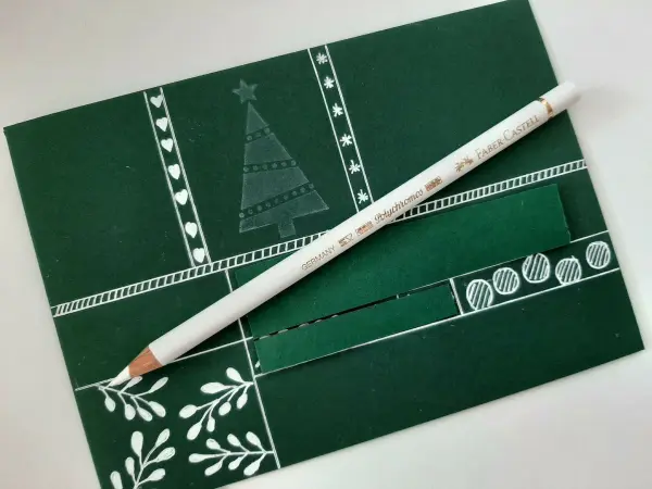 Ein gefalteter Tannenbaum aus grüner Karte liegt neben einem weißen Farbstift auf einer dekorativen Karte im Weihnachtsdesign.