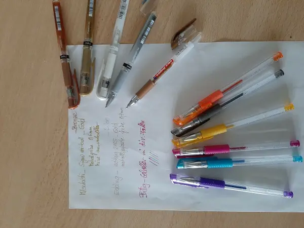 Auf einem Tisch liegen verschiedene bunte Stifte und ein Blatt Papier, auf dem weiße und goldene Tinte notiert ist.