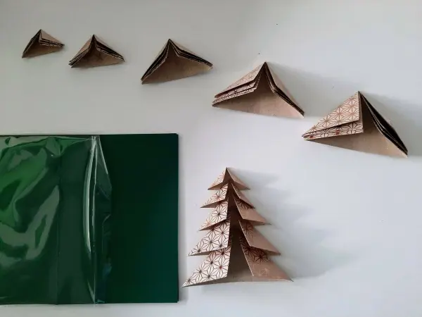 Ein gefalteter Tannenbaum steht in der Mitte, umgeben von kleineren gefalteten Formen und einer grünen Kartenhülle.