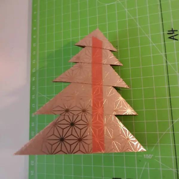 Ein gefalteter Tannenbaum aus glänzendem Papier steht auf einem grünen Schneideunterlage, dekoriert mit geometrischen Mustern.