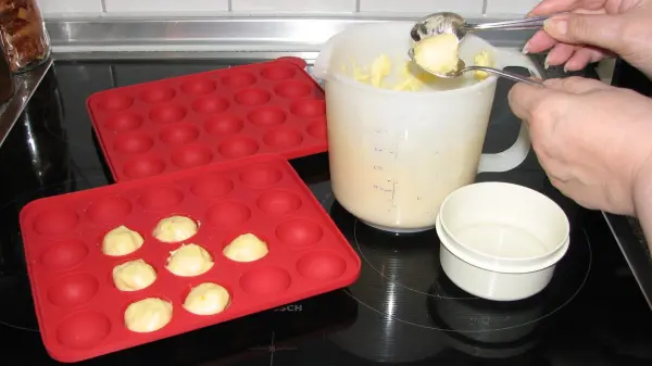 Eine Person formt Frischkäse-Schneebällchen mit einem Löffel, während leere Silikonformen auf einer Küchenplatte liegen.