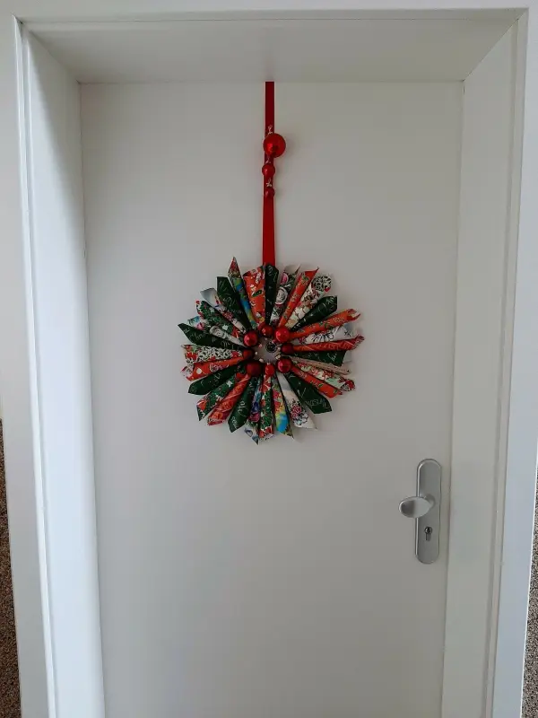 Ein bunter Weihnachtskranz aus Papierrollen und Tannenbaumkugeln hängt dekorativ an einer weißen Tür.
