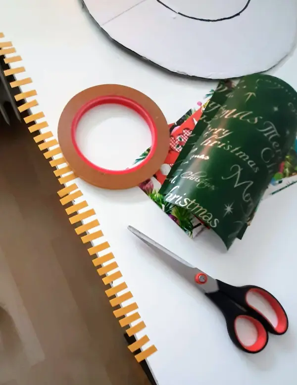 Eine Rolle mit weihnachtlichem Papier und eine Schere liegen auf einem Tisch, während ein Weihnachtskranz hergestellt wird.