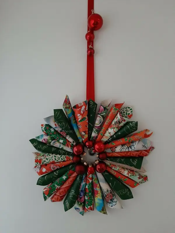 Ein bunter Weihnachtskranz aus Papierrollen und roten Kugeln hängt an einem roten Band an der Wand.