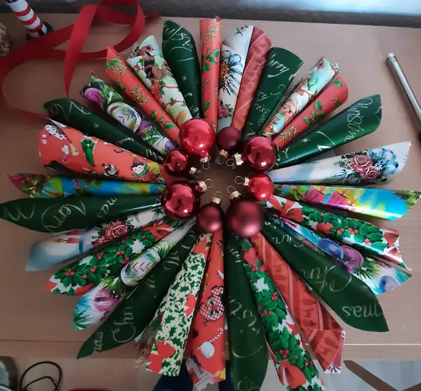 Ein bunter Weihnachtskranz aus Papierrollen und roten Tannenbaumkugeln liegt auf einem Tisch und strahlt festliche Stimmung aus.