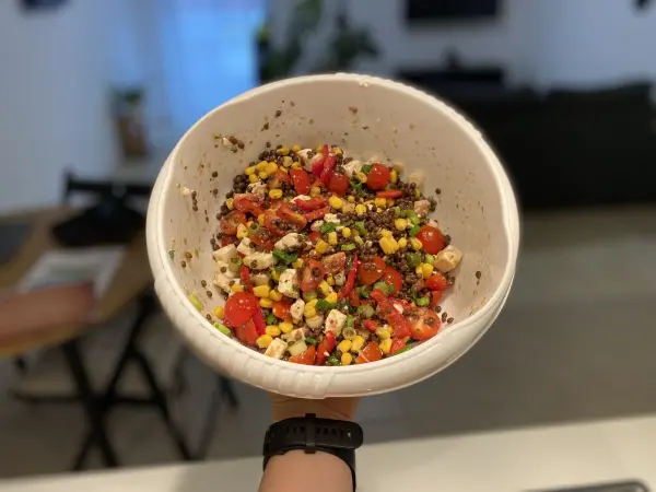 Eine Person hält eine weiße Schüssel mit einem bunten Salat aus Belugalinsen und Mais in einer modernen Küche.