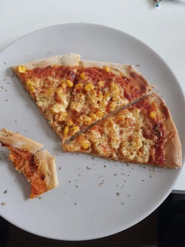 Eine Pizza mit Käse und Mais liegt auf einem weißen Teller, während ein Stück davon abgebissen wurde.