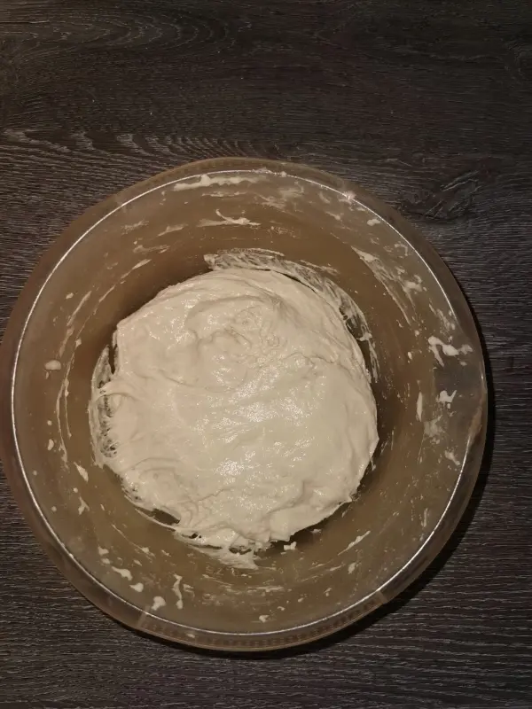 Ein runder Pizzateig ruht in einer transparenten Schüssel auf einem dunklen Holzuntergrund.