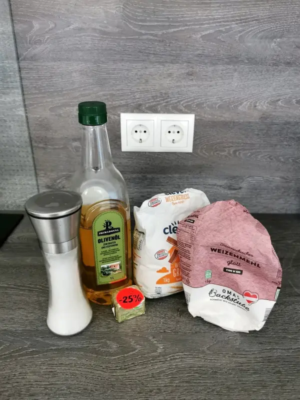 Auf einem Küchentisch liegen Zutaten für Pizzateig: Mehl, Olivenöl, Salz und Butter. Eine moderne Umgebung umrahmt das Bild.