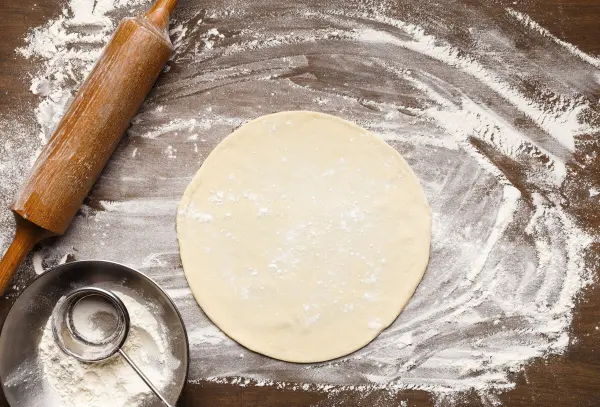 Ein runder Pizzateig liegt ausgerollt auf einer bemehlten Holzfläche neben einem Nudelholz und einem Sieb.