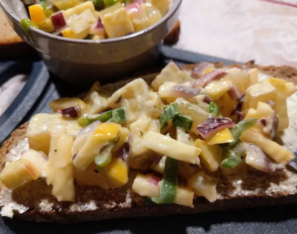 Auf einem Stück Brot liegt ein Handkäse-Tatar mit bunten Würfeln, umgeben von einem kleinen Schälchen mit der restlichen Mischung.