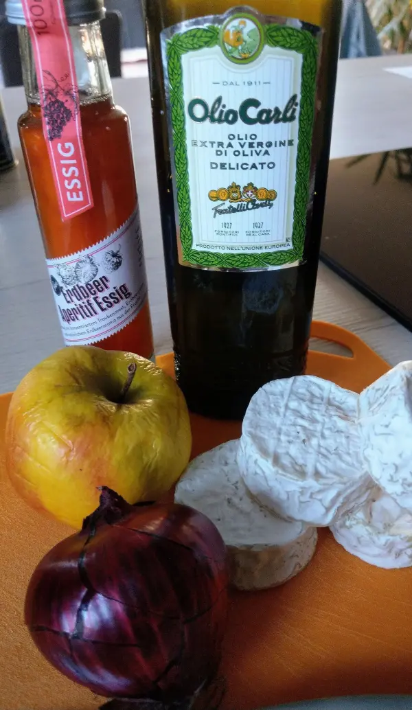 Auf einem orangefarbenen Schneidebrett liegen ein Apfel, Zwiebeln und frischer Handkäse, umgeben von Essig und Olivenöl.