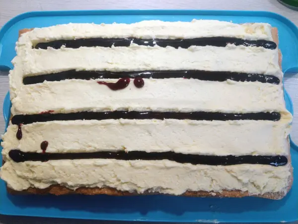Ein rechteckiger Frankfurter Kranz, mit Creme und Schokoladensoße, liegt auf einem blauen Teller in einer hellen Küche.