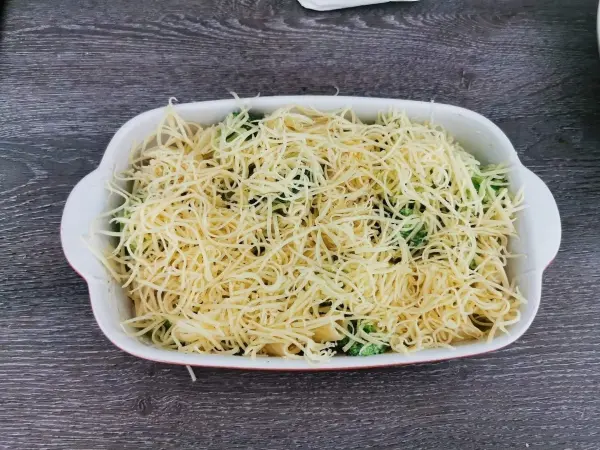 Eine Auflaufform mit Tortiglioni und Brokkoli, bedeckt mit einer Schicht geriebenem Käse, steht auf einem Holztisch.