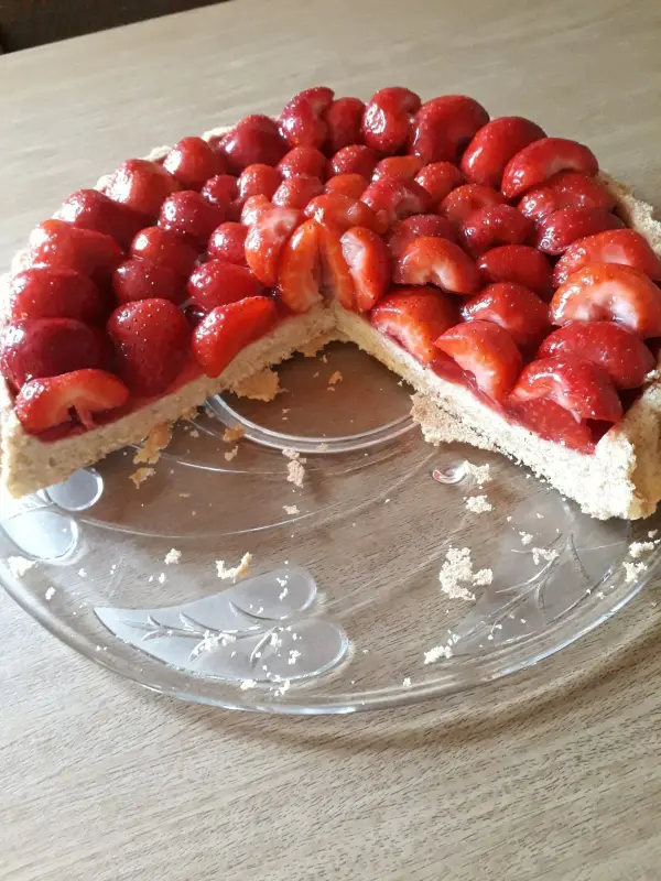 Ein Erdbeerkuchen mit Dinkeltortenboden steht auf einem Teller, mit einer Stück davon herausgeschnitten, umgeben von einer Holzfläche.